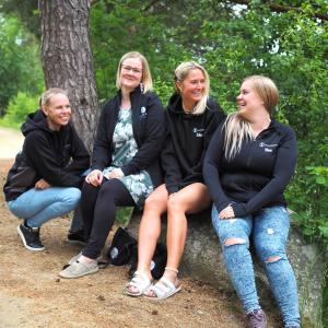 Nuorisotoimen työntekijät Sanna Tuominen, Jennika Salonen, Erika Raitanen ja Nina Kuusisto-Huuskonen istuvat hymyilleen puun rungon päällä. Taustalla vihreän kesäinen metsämaisema.