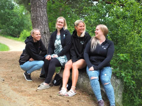 Nuorisotoimen työntekijät Sanna Tuominen, Jennika Salonen, Erika Raitanen ja Nina Kuusisto-Huuskonen istuvat hymyilleen puun rungon päällä. Taustalla vihreän kesäinen metsämaisema.