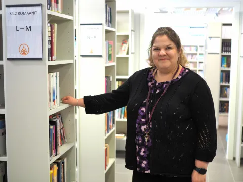 Kirjastotoimenjohtaja Salla Mäkinen katsoo hymyillen kameraan. Taustalla kirjaston hyllyjä.
